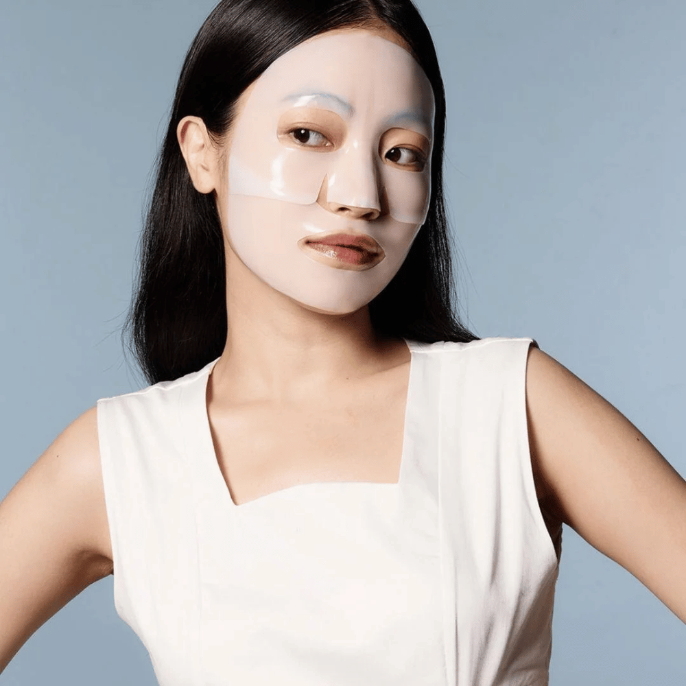 Una mujer con un vestido blanco sin mangas lleva una mascarilla facial transparente que cubre casi todo su rostro. La máscara da una impresión de cuidado avanzado de la piel y proporciona una apariencia de tratamiento de belleza, diseñada para hidratar profundamente y tratar la piel.